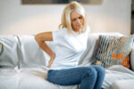 Upset frustrated mature woman suffering from backache, unhappy senior blonde sitting on a sofa at living room, feeling discomfort because of pain in back, have joint problems, need rest